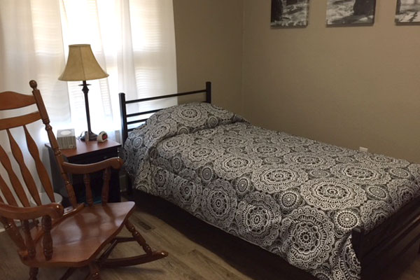 A twin-sized bed with floral sheets and wooden rocking chair in a resident's bedroom inside MHAST's Crisis Respite Supportive Residence